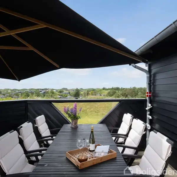 Sort spisebord på terrasse med otte hvide stole, placeret under stor parasol. Der står blomstervase og serveringsbakke med vin og glas på bordet. Grøn landskab baggrund.