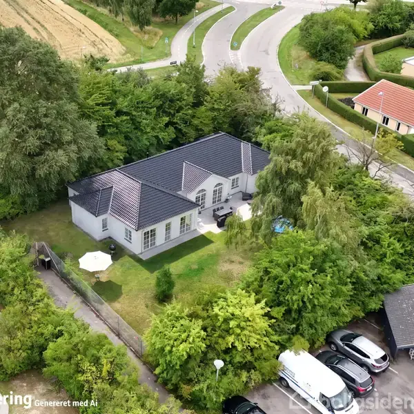 Et hus med mørkt tag ligger omgivet af træer og grønne områder. Foran huset er der en terrasse med møbler og en parasol. Stier og veje omkranser ejendommen.