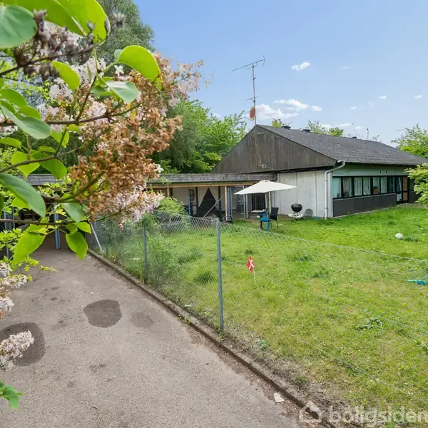 En villa ligger i en grøn have med en grusvej og blomstrende buske i forgrunden. Haven er indhegnet og har en terrasse med parasol. Himmel er klar.