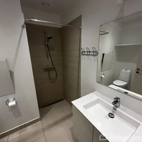 Spejl over hvid vask med skuffe. Bruser i brusekabine til venstre, flisevæg. Toilet med rulleholder i baggrunden. Neutrale farver, lysflisegulv.