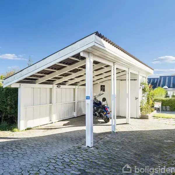 En hvid carport med fladt tag rummer en motorcykel. Omgivet af hække og en brostensbelagt indkørsel ved siden af en murstensbygning under en klar blå himmel.