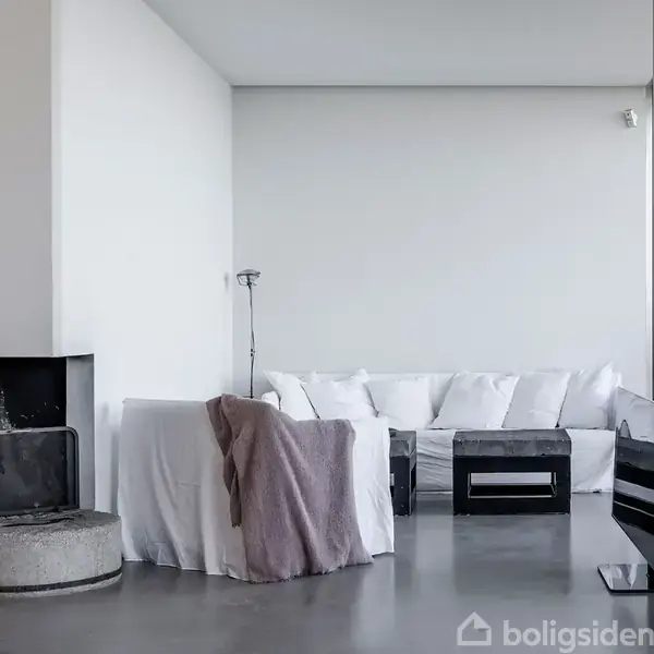 Sofa med hvide puder står i en minimalistisk stue. En lænestol er dækket af et tæppe ved siden af. Rummet har grå cementgulve og en moderne pejs.