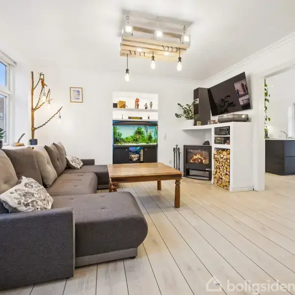 En grå sofa står i en lys stue med trægulv. Et stort akvarium på en reol er i fokus, omgivet af planter og dekorationer. I baggrunden ses et køkkenområde.