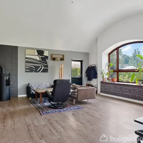 Stue med moderne indretning inkluderer sofaer og lænestol omkring sofabord på gulvtæppe; store vinduer med udsigt til natur; hylder med planter og bogreol til venstre for optisk balance.