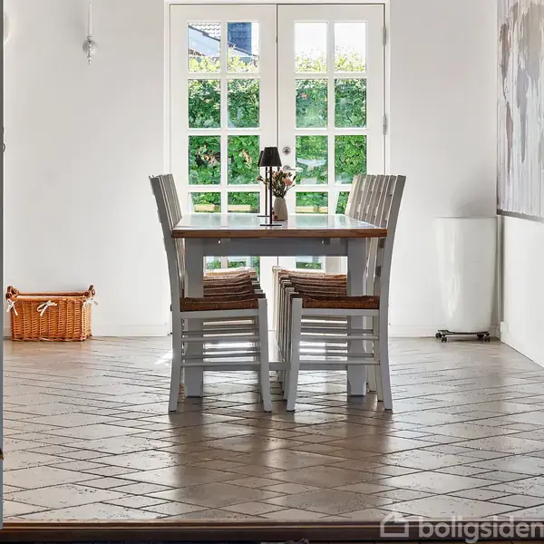 Et træbord med fire stole står på et flisegulv i et lyst rum. Store glasdøre bagved åbner ud mod en grøn have. En kurv står i hjørnet.