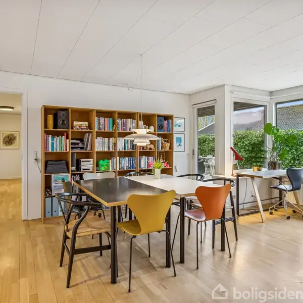 En spisestue med et stort bord omgivet af farverige stole; en reol fuld af bøger står op ad væggen. Store vinduer giver udsigt til en grøn hæk udenfor.