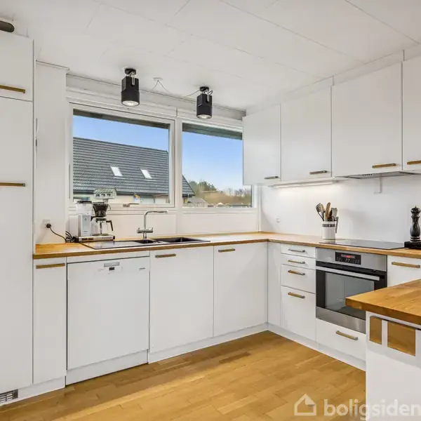 Et moderne køkken har hvide skabe og træbordplader. Køkkenet omfatter en ovn, vask, og kaffemaskine med udsigt til et vindue, der viser tagene af nærliggende bygninger.