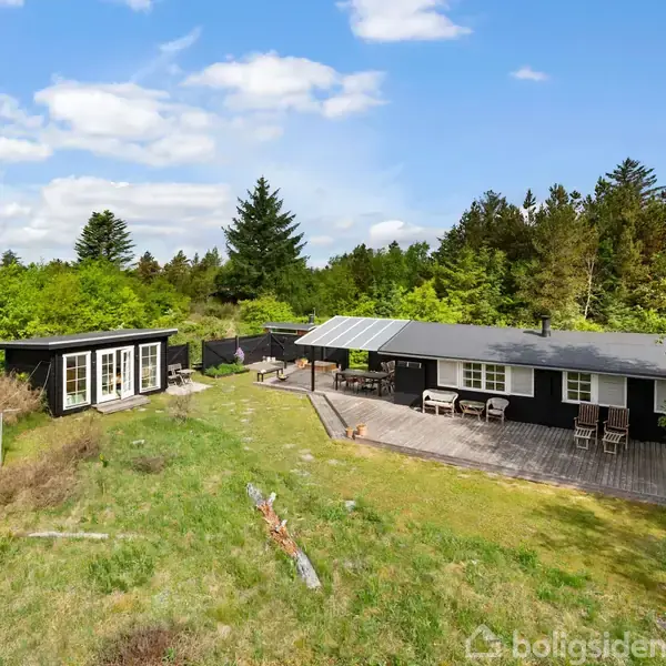 Sommerhus i sort træ med terrasse og havemøbler, omgivet af grønne træer og buske. Til venstre står et mindre sort anneks med glasdøre. Blå himmel med få skyer.