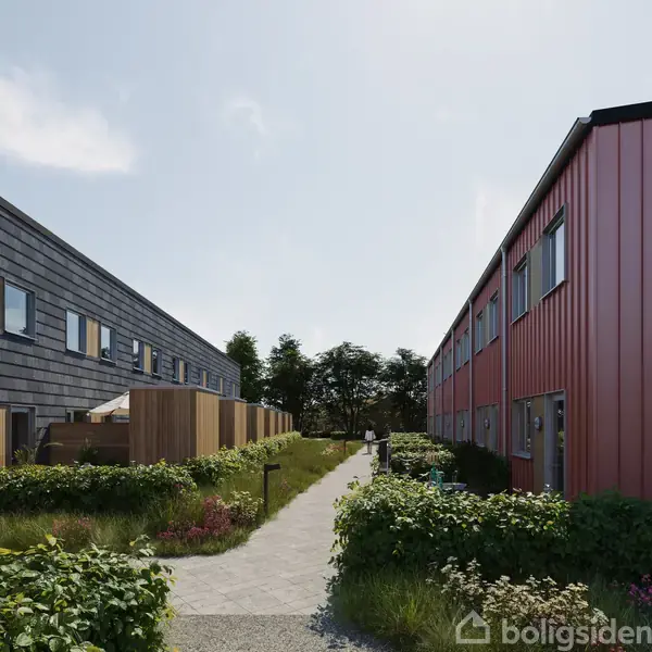 To rækker af moderne rækkehuse i grå og rød farve omgiver en smal sti med grønne buske, der løber mellem dem, med træer i baggrunden under en klar himmel.