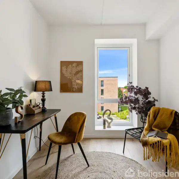 Et stilfuldt, moderne kontor har en sort skrivebord med planter og lampe, en gylden stol, en plakat på væggen, og et stort vindue med udsigt til et byggeri udenfor.