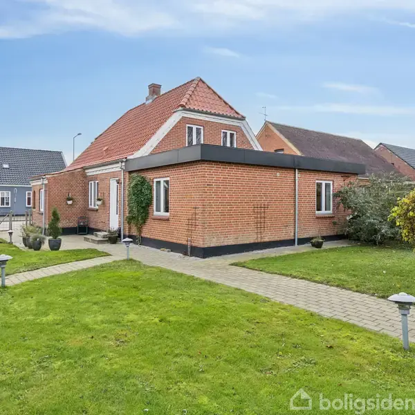 En murstenshus står stille, omgivet af en velplejet grøn have med en flisebelagt sti. Nabohuse med lignende arkitektur ses i baggrunden under en klar, blå himmel.