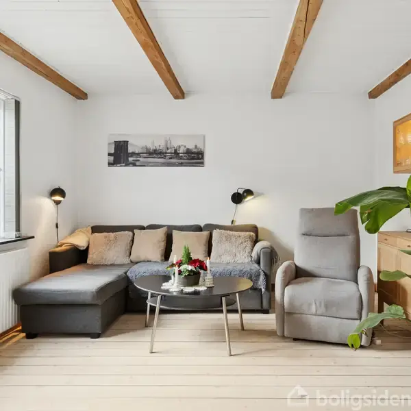 En stue med en hjørnesofa og lænestol vendt mod et rundt sofabord. Væggene har en hylde med glaslåger. Store vinduer lader naturligt lys strømme ind.