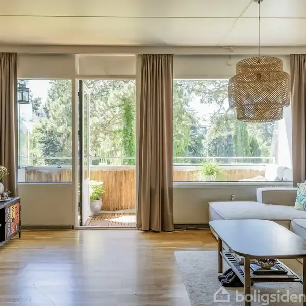 Stue med beige sofa og træbord, fladskærms-TV på væggen, åbne døre fører til balkon med udsigt over grønne træer; trælaminatgulv og hængende lampe.
