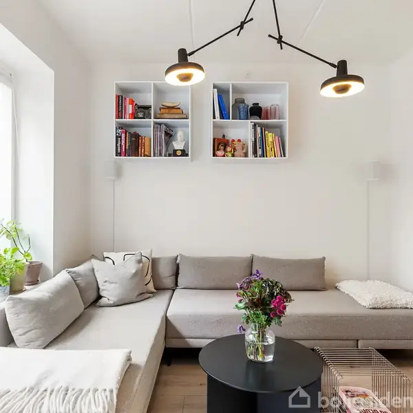Gråt sofaarrangement står i stuehjørne. Sofabord med buket i midten. På væggen er hylder med bøger og dekorationer. Store vinduer med planter skaber et lyst miljø.