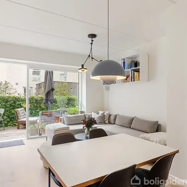 Spisebord med fire stole står i en lys stue, hvor en hjørnesofa med puder og væghylde ses. Glasdør fører ud til terrasse med møbler og grønne omgivelser.