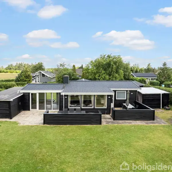 Et sort-hvidt sommerhus står stille på en velplejet grøn plæne. Store vinduer vender mod en flisebelagt terrasse. Omgivet af grøn beplantning og nabohuse i en landlig setting.