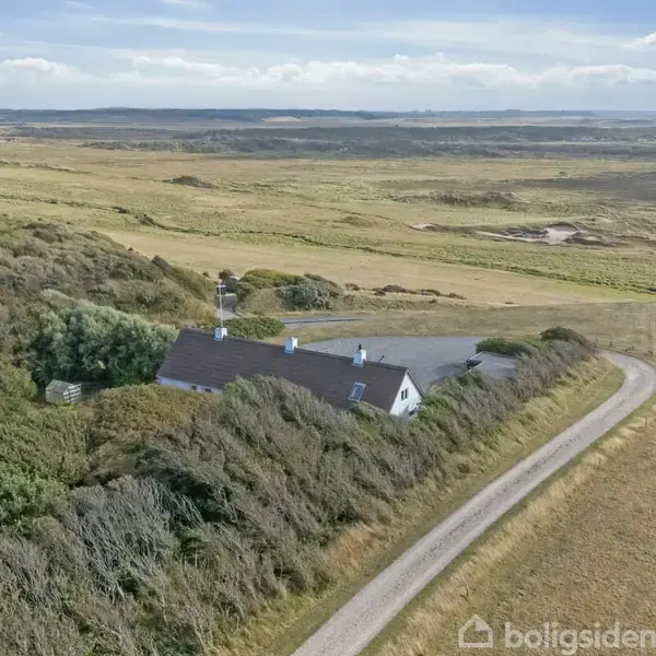 Huset ligger blandt træer og buske. Det er en hvid bygning med sort tag, placeret ved en snoet grusvej. Omgivelserne er fladt landbrugslandskab og åbne marker.