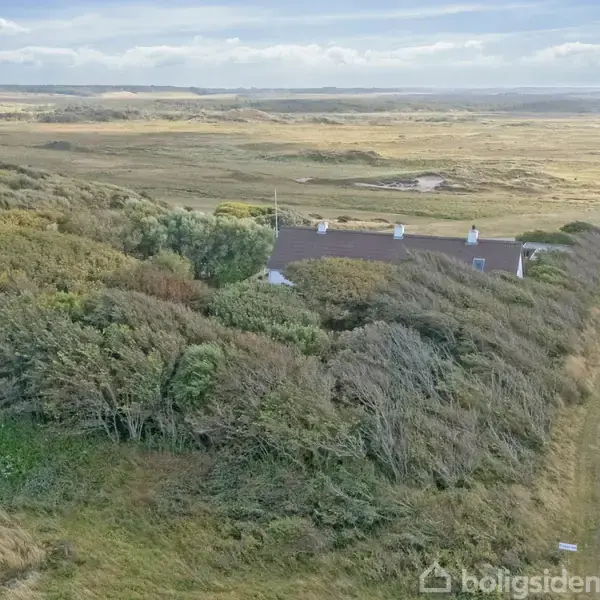 En hvid bygning med mørkt tag ligger skjult blandt tætte træer. En snoet grusvej løber forbi til højre. Landskabet er fladt og åbent med græsarealer i baggrunden.