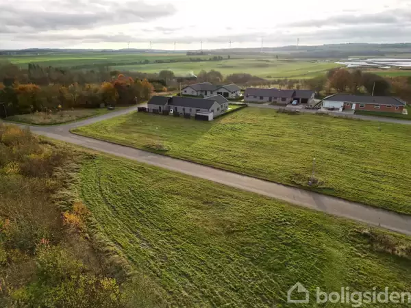 Huse står stille blandt marker og træer; asfalteret vej løber forbi. Vindmøller kan skimtes i baggrunden under en overskyet himmel.