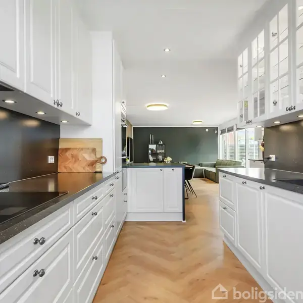Hvidt køkken strækker sig i et langsgående layout med skinnende sorte bordplader, tilstødende en åben stue med spisebord og stilfuldt sofahjørne under indbyggede loftslamper.