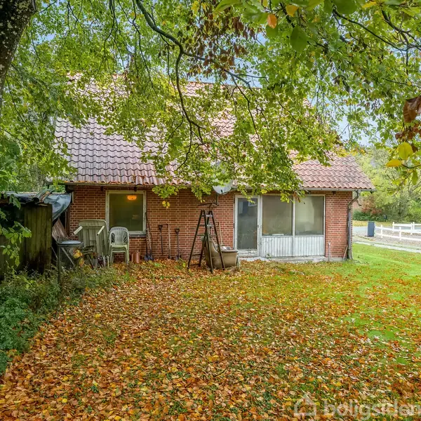 Et rødt murstenshus omgivet af efterårsblade ligger ved en græsplæne. Husets gavl har store vinduer, og der står havemøbler ved siden af en stige under et stort træ.