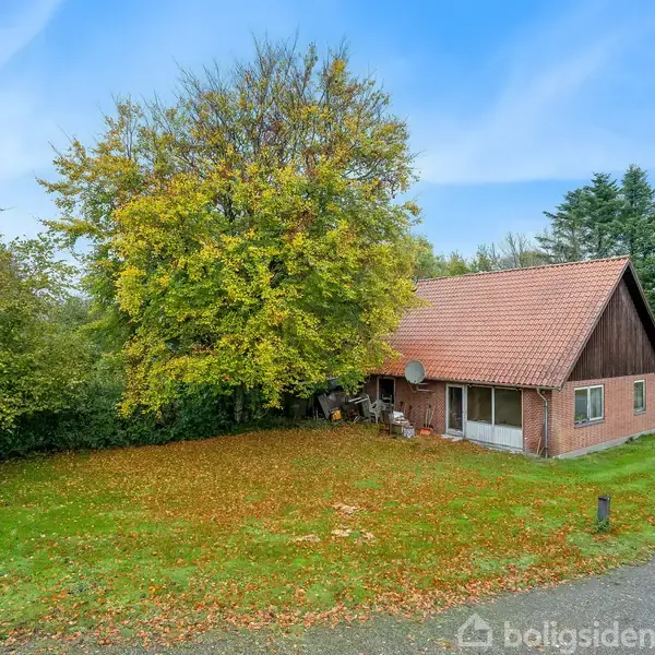 En rød murstensbygning med rødt tegltag står ved en grusvej og er omgivet af grønne træer. Blade dækker jorden foran huset, hvilket indikerer efterår.