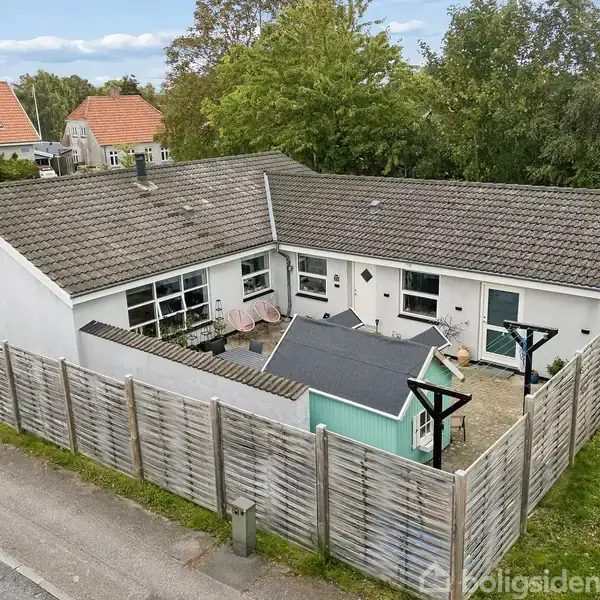 Et hvidt enfamiliehus med fladt tag, omgivet af et træhegn. På terrassen står to lyserøde stole ved et lille bord, og der er en mintgrøn skur.