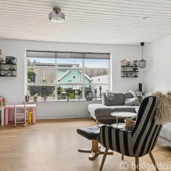 Stue med grå sofa, sort-hvid-stribet lænestol, og fladskærms-TV. Store vinduer med udsigt til have, indrettet med legetøj og reoler. Lyse trægulve og hvidt loft med lampe.