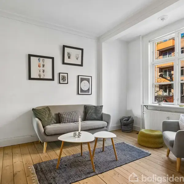 Sofa står i en lys stue, flankeret af et sofabord med stearinlys. Billeder hænger på væggen over sofaen. Et vindue giver udsigt til en murstensbygning.