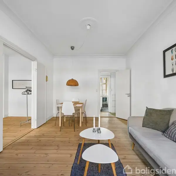 Sofa står i stue med sofabord, set fra dør. Lys træbeklædning på gulv, spisebord med stole i baggrunden. Kunst hænger på væggen, minimal indretning, hvidmalede vægge.