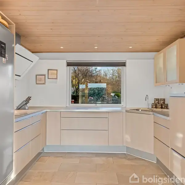 Et moderne køkken med lyse skabe og stålapparater. Rummet har et stort vindue, der giver udsigt til en have, og træplankeloft, der skaber en lys og indbydende atmosfære.