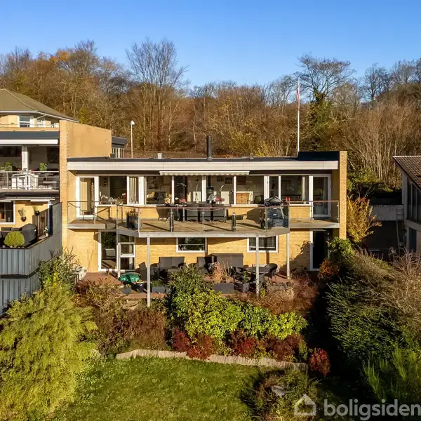 Et moderne to-etagers hus med store vinduer, som står på en skrånende grund omgivet af frodig beplantning og naboer. Terrasse på både øverste og nederste etage.