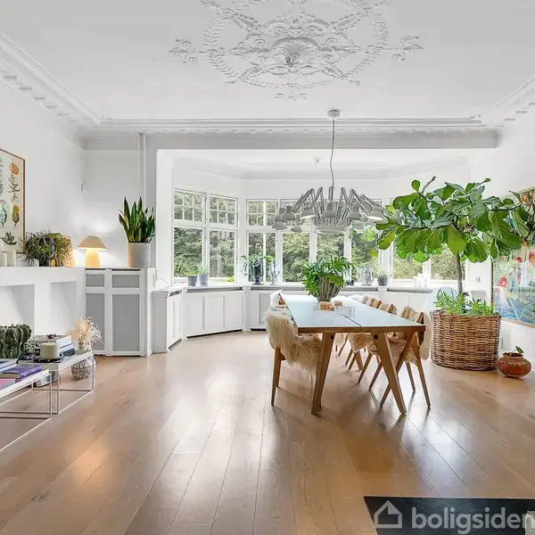 Et spisebord står midt i et lyst rum med trægulve. Omkring bordet er der stole, store grønne planter og kunst på væggene. Vinduer fylder bagvæggen.