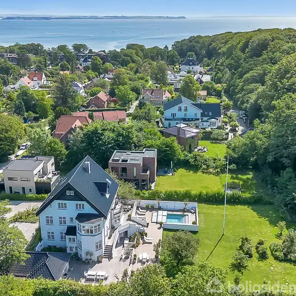 Stort hus med sort tag og tilstødende terrasse samt pool, beliggende i en grøn, beboet landsby nær en skov. Haven er velholdt med en flagstang. Havet kan ses i baggrunden.