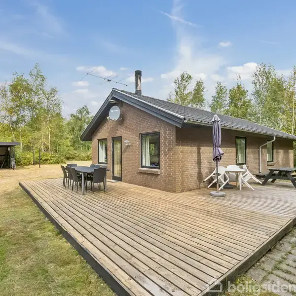 Et træhus står stille på en stor, græsbeklædt grund. Omkring huset er der skov og en stor træterrasse med udendørs møbler. En carport ses i baggrunden.