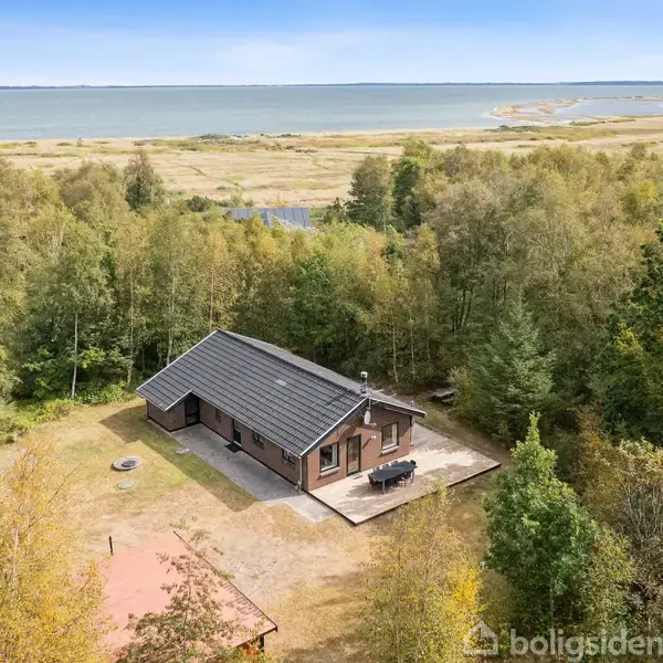 Et træhus står stille omgivet af tæt skov med en terrasse. I baggrunden ses en smuk udsigt over en bred sø eller fjord, under en klar blå himmel.