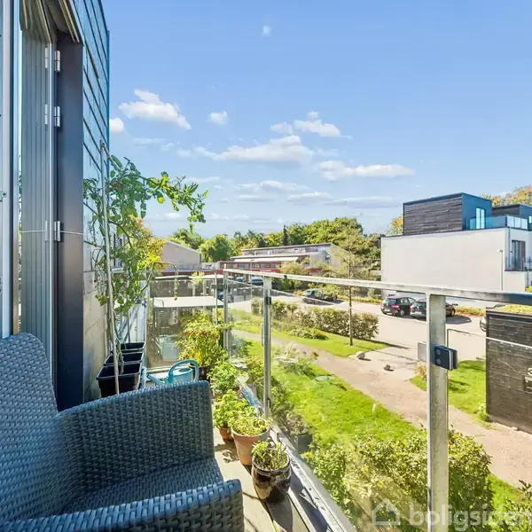 En balkon med en fletstol står ved siden af en glasbalustrade, omgivet af planter og med udsigt til en moderne boligområde på en solrig dag.