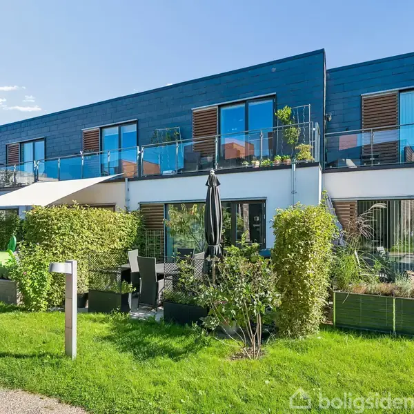 Rækkehuse med blå facader og store vinduer. Foran er en terrasse med møbler og planter. Græsplæne og sti i forgrunden. Solrig dag med klare himmel.