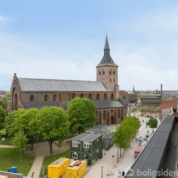 En kirke med et højt spir står blandt grønne træer i en byscene. En moderne bygning med en balkon ses til højre i billedet.