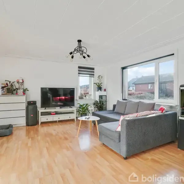 Et moderne, lyst stue med trægulv. Sofa og sofabord står foran et stort vindue. TV står på et skab, og der er et akvarium til højre. Indretningen er minimalistisk.