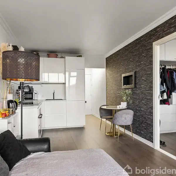 En sofa med puder står i en moderne, lille stue. Ved siden af er et kompakt køkken med hvide skabe. Et lille spiseområde har to stole ved et rundt bord.