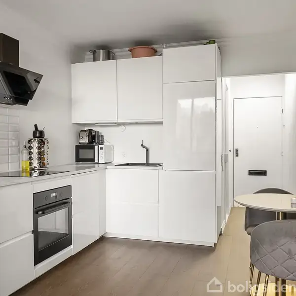 Køkken med hvide skabe og apparater, herunder ovn og mikroovn. En lille spiseplads med rundt bord og to stole står til højre, i en moderne boligindretning.