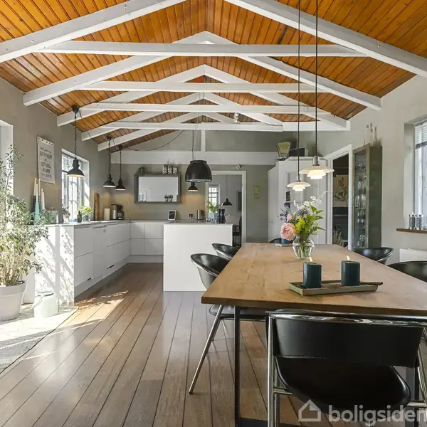 Et lyst køkken-alrum med træloft og synlige bjælker. Langbord i midten omgivet af stole, planter og moderne lamper. Store vinduer slipper naturligt lys ind.