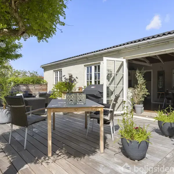 Træbord med stole står på en træterrasse foran et hvidt murstenshus. Døre står åbne mod haven, og krukker med planter er placeret rundt omkring. Sollys skinner ned.