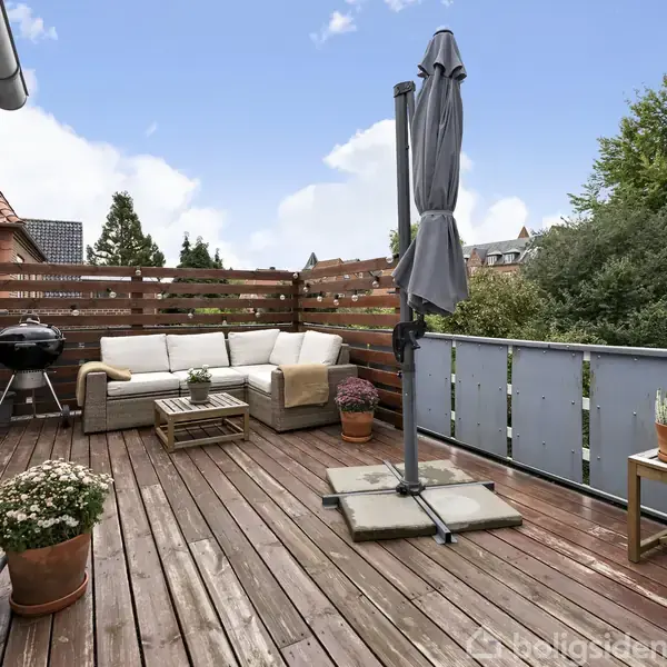 En terrasse med træbrædder har en hjørnesofa og et havebord, omgivet af en træhegn. En grill står til venstre, og planter er i krukker. Himlen er klar.