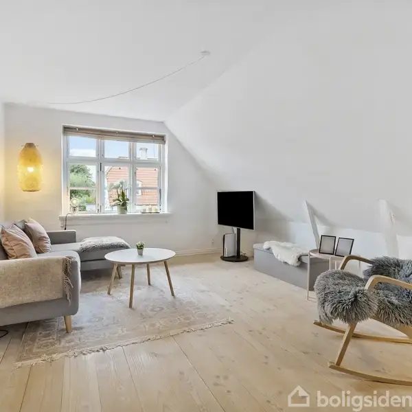 En grå sofa står i en lys stue med trægulv, flankeret af en lænestol med pels, en lampe og et lille bord, med et stort vindue og tv i baggrunden.