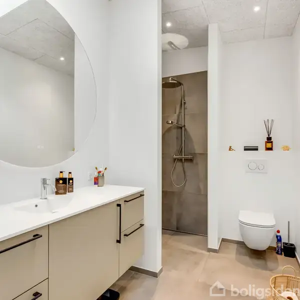 Rundt spejl hænger over et moderne badeværelsesskab med håndvask, omgivet af tandbørster og sæber. Til højre er et toilet, ved siden af en stilfuld brusekabine.