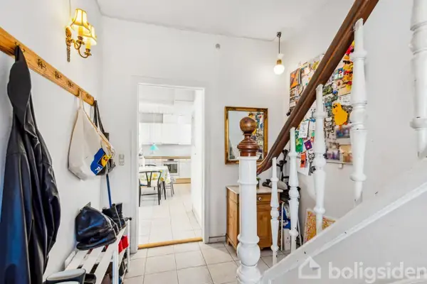 Trappe fører opad i indgangsparti med frakkekrog til venstre og spejl til højre. Døråbning fører til køkken med synlige stole og bord i baggrunden.