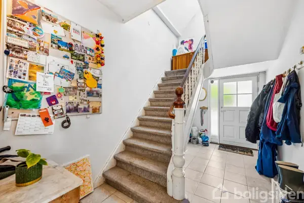 Trappeopgang beklædt med tæppe fører opad; væg til venstre dekoreret med opslagstavle fyldt med billeder og noter; entréen har frakkeophæng og flisegulv ved siden af hoveddør.