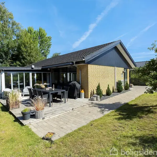 Et gult murstenshus med sort tag ses fra havevinklen, med havemøbler på en flisebelagt terrasse omgivet af græsplæne og buske under en klar blå himmel.
